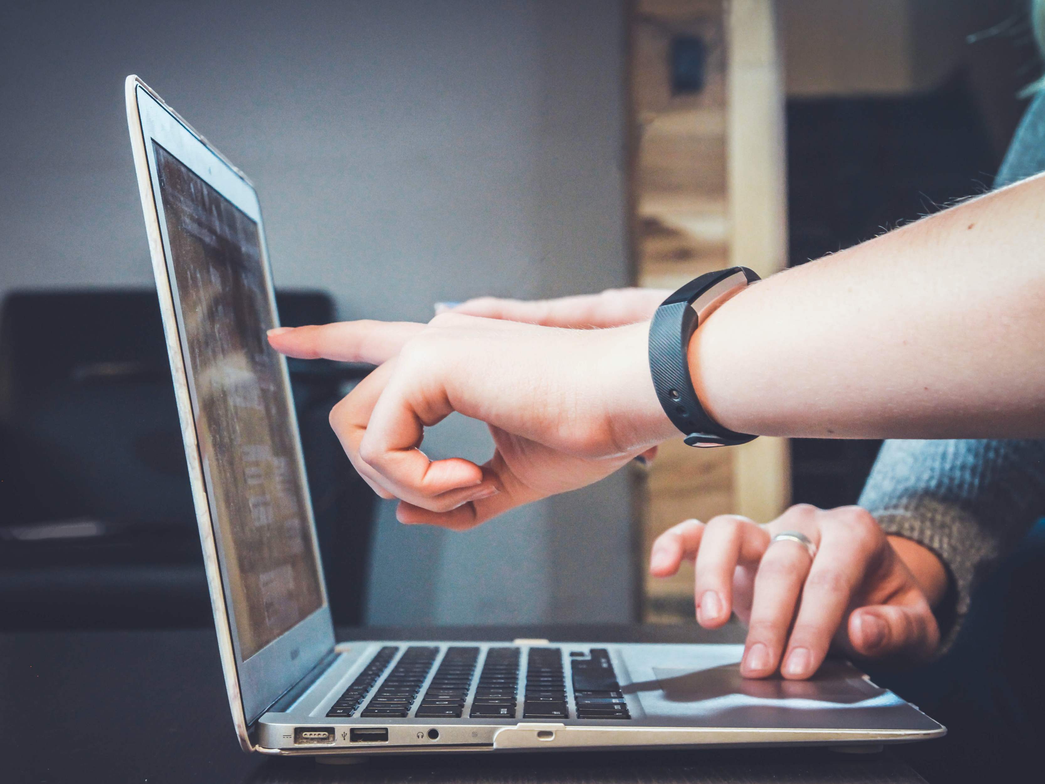 A laptop is open with one person using the trackpad mouse and one person pointing to the screen